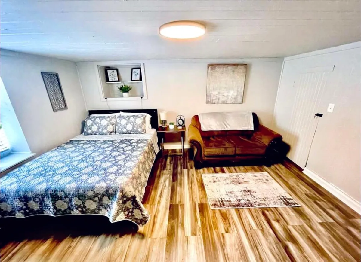 A cozy bedroom scene with a bed adorned in floral bedding, a comfortable brown couch adjacent to the bed, and decorative artwork on the walls. Natural light streams gently into the room, highlighting the inviting, restful ambiance and emphasizing soft, tranquil lighting.