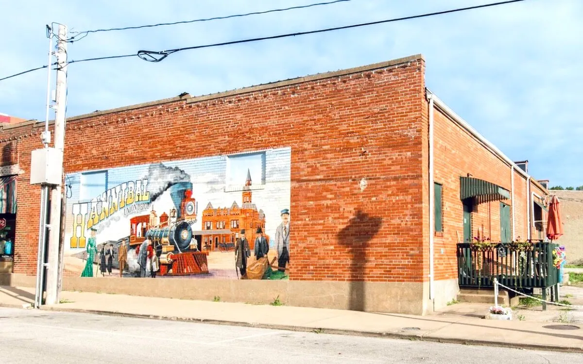 A large, vibrant mural depicting a steam train and historical figures on a brick wall, with outdoor seating and umbrellas in the foreground. The setting is lively and artistic, capturing a unique aspect of Hannibal's cultural identity.