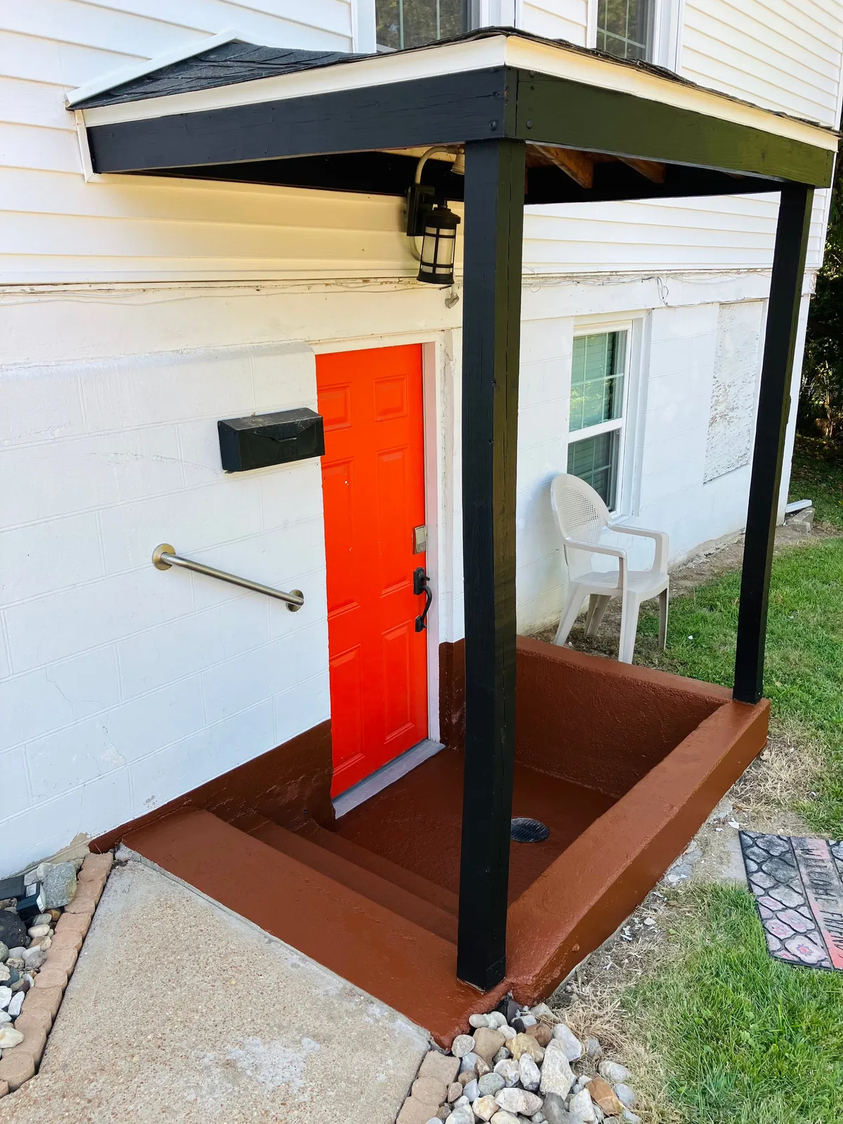 The welcoming exterior of Huck's Modern Hideaway, highlighted by a bright orange door and a sleek black awning. The front porch area features comfortable outdoor seating, suggesting a friendly and accessible home base for guests.