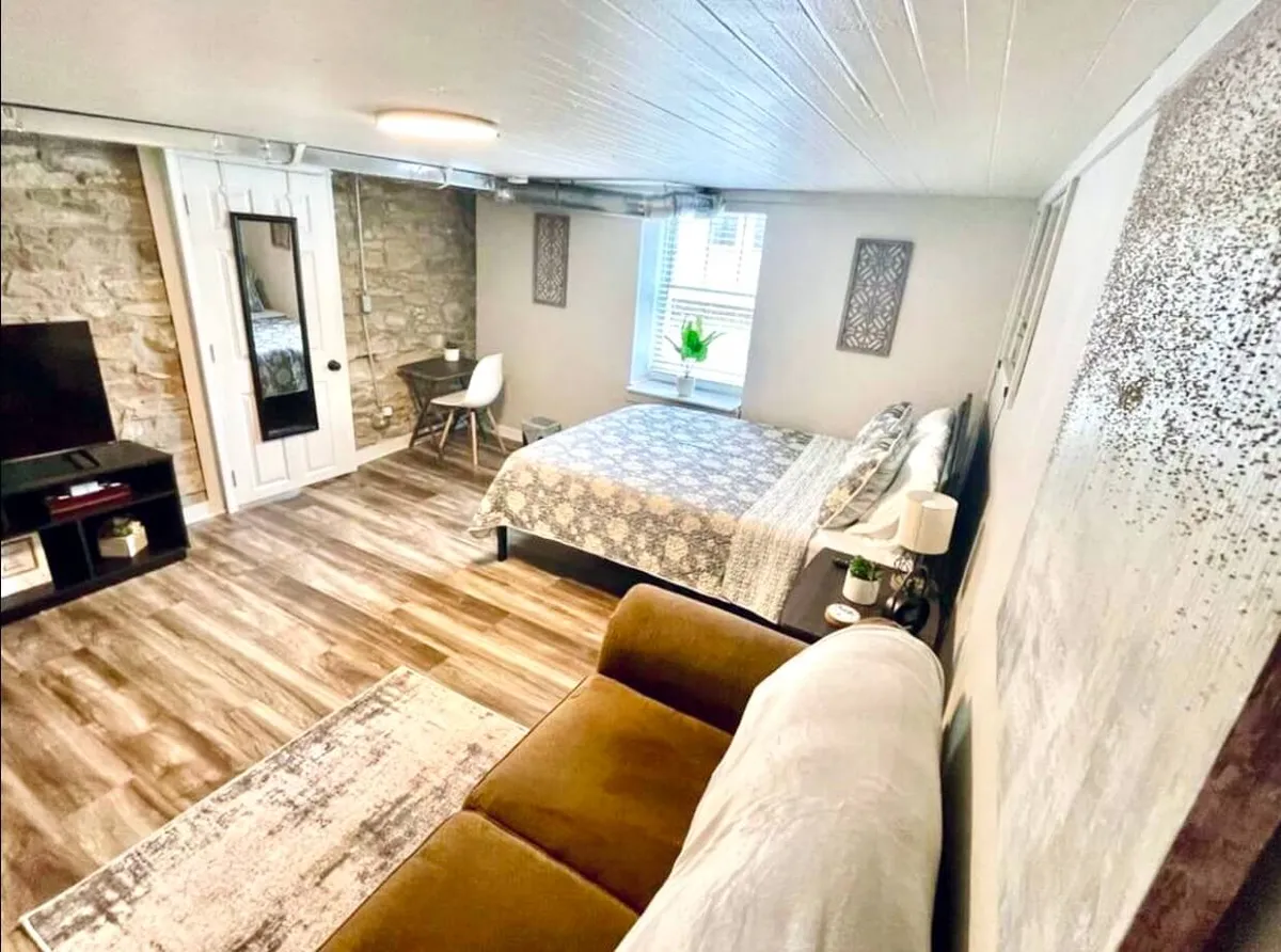 The image shows a living space with a bed dressed in decorative bedding, a plush sofa, a stone accent wall, a window with a plant, and a TV stand. The setting is warm and inviting, hinting at relaxing movie nights, quiet mornings, and cozy reading afternoons.