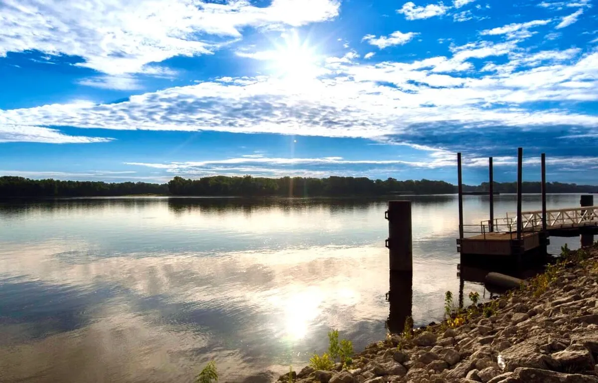 A peaceful view of the river, with sunlight sparkling on the water, a small dock structure, and a rocky shoreline. The scene radiates tranquility and hints at outdoor activities such as boating or riverside picnics.