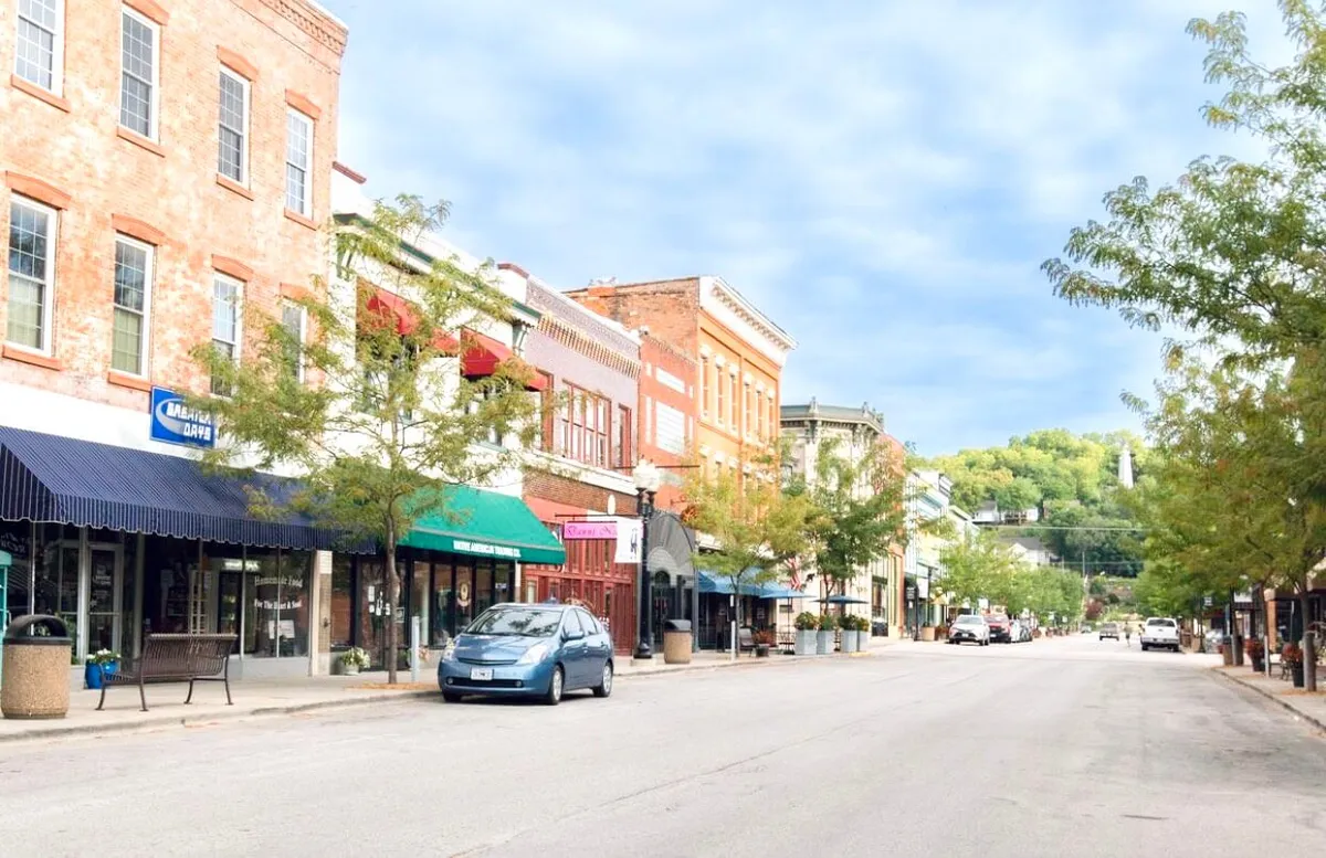 A lively Hannibal streetscape featuring historic brick buildings, colorful shopfront awnings, leafy trees, and parked cars. People can be seen enjoying outdoor cafes, shopping, and strolling along the sidewalks, evoking the energy and community charm of festival days.