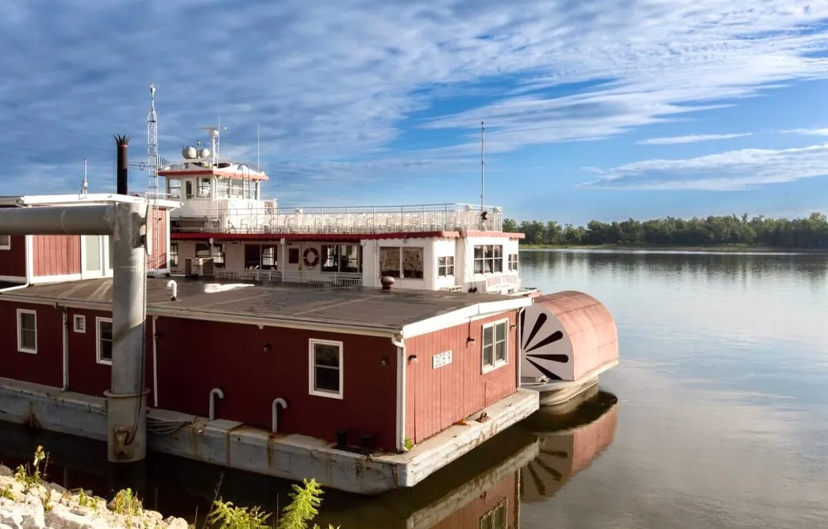 A houseboat moored on the river with an inviting deck area, open sky, and water views. The deck is set up for relaxing or dining outdoors, emphasizing the proximity and connection to the river.